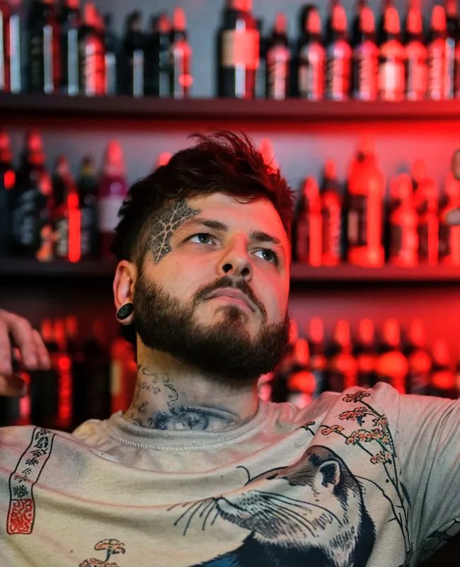 Alexander Kuzmin, tattooed face and neck, sitting in front of a red-lit wall of tattoo ink bottles at Requiem Tattoo Studio