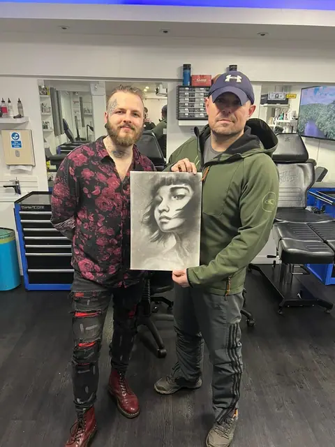 two men holding a black and grey female portrait drawing in a tattoo studio
