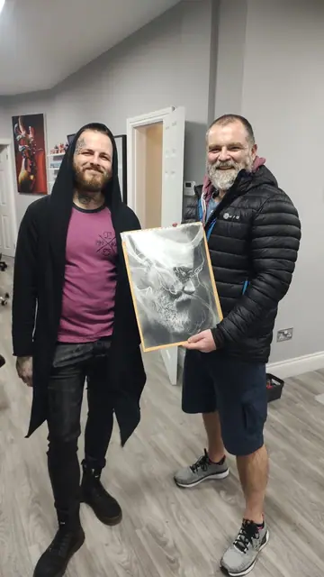 two men holding black and white framed artwork, one man has face tattoo